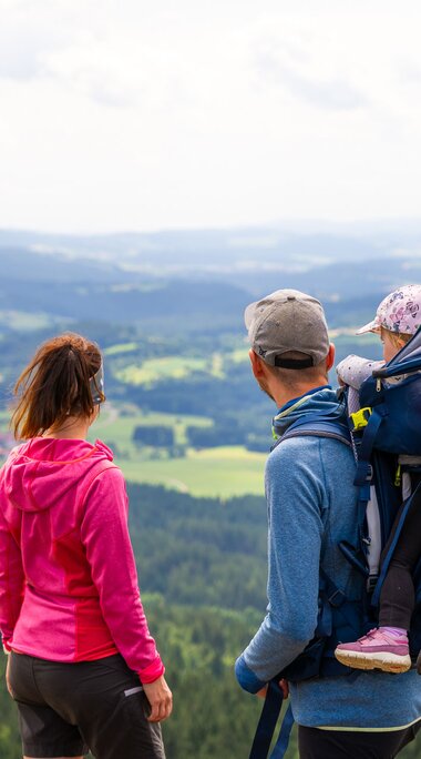 Ein Paar steht zusammen mit einem Kleinkind in der Kindertrage auf dem Rücken des Vaters auf einem Gipfel und blickt in die Ferne. | © Bodenmais Tourismus & Marketing GmbH