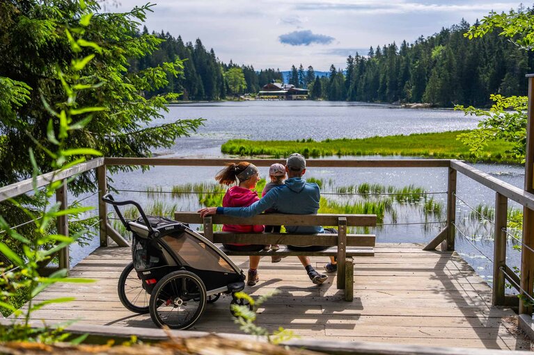 Mutter, Vater und Kleinkind sitzen auf einer Bank mit Blick auf den Großer Arbersee sowie das Arberseehaus | © Bodenmais Tourismus & Marketing GmbH