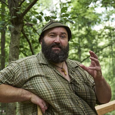 Der Woid Woife, ein großer, kräftiger Mann mit Filzhut und kariertem Hemd, steht im Wald, hat die Hand in die Hüfte gestemmt und erklärt etwas. | © Bodenmais Tourismus & Marketing GmbH