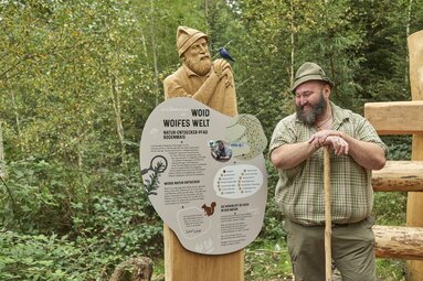 Der Woid Woife steht am Schild des Natur-Entdecker-Pfades, in dem er im Holz abgebildet ist und lacht. | © Bodenmais Tourismus & Marketing GmbH