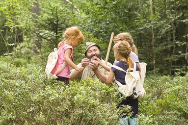Der Woid Woife hält etwas in der Hand was die drei Mädchen um ihn herum betrachten. | © Bodenmais Tourismus & Marketing GmbH