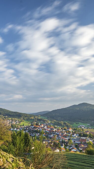 Hinter Bäumen und Hügeln versteckt liegt Bodenmais. Vor der Ortschaft ist eine gemähte Wiese zu erkennen. Am blauen Himmel zeichnen sich weißen Wolken ab. | © Bodenmais Tourismus & Marketing GmbH