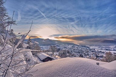 Über glitzernden Schnee hinweg blickt man auf Bodenmais. Über der Ortschaft wacht der Silberberg, hinter dem die Sonne hervor blitzt, der blaue Himmel ist durchzogen von Schleierwolken. | © Bodenmais Tourismus & Marketing GmbH