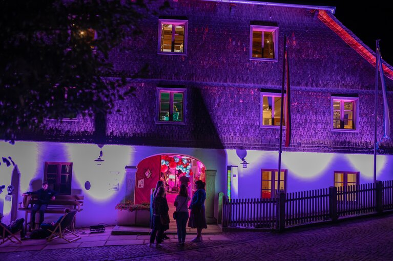 Das Alte Rathaus wird in lila Licht gehüllt. Die Eingangstür steht offen, davor stehen Menschen und unterhalten sich. | © Bodenmais Tourismus & Marketing GmbH