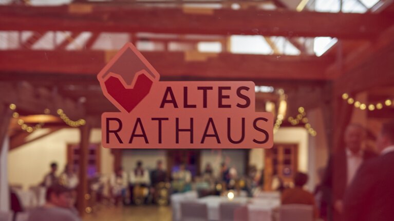 Eine Glastür ist mit der Aufschrift "Altes Rathaus" und dem Bodenmais Logo versehen. Im Raum dahinter sieht man Lichterketten, weiße Hussen und Menschen. | © Bodenmais Tourismus & Marketing GmbH