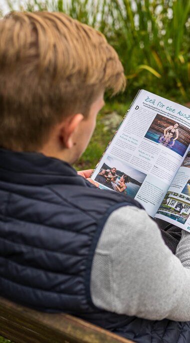 Der Blickt fällt über die Schulter eines Mannes, der auf einer Bank sitzt und eine Zeitschrift liest. | © Bodenmais Tourismus & Marketing GmbH
