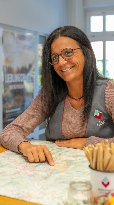 Eine Frau mit dunklen, langen Haaren und Weste, auf der der Name "Elke" steht, berät eine Frau am Tresen und zeigt mit dem Finger auf einen Ortsplan. | © Bodenmais Tourismus & Marketing GmbH