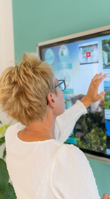 Eine Frau mit kurzen Haaren bedient mit der Hand ein Display, auf dem verschiedene Attraktion angezeigt werden. | © Bodenmais Tourismus & Marketing GmbH