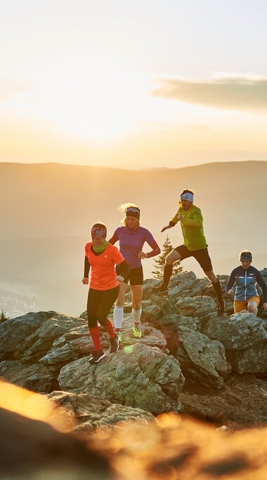 Vier Trailrunner laufen über einen steinigen Gipfel. Unter ihnen liegt Bodenmais, bereits im Schatten der untergehenden Sonne. | © Bodenmais Tourismus & Marketing GmbH
