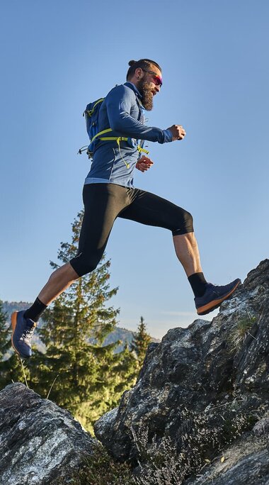 Ein männlicher Trailrunner springt auf einen Stein. Ein Bein ist dabei noch in der Luft. Hinter ihm ist Wald. | © Bodenmais Tourismus & Marketing GmbH