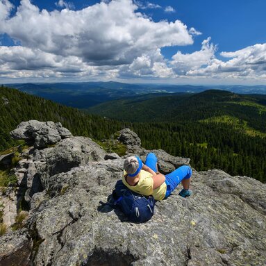 Eine Frau in sportlicher Kleidung liegt bequem auf ihren Rucksack zurückgelehnt auf einem Gipfel und schaut in die Ferne. Um den Gipfel stehen grüne Wälder. | © Bodenmais Tourismus & Marketing GmbH