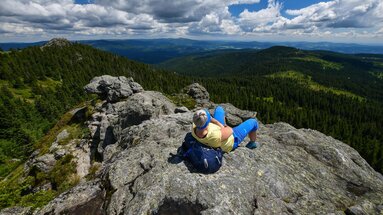 Eine Frau in sportlicher Kleidung liegt bequem auf ihren Rucksack zurückgelehnt auf einem Gipfel und schaut in die Ferne. Um den Gipfel stehen grüne Wälder. | © Bodenmais Tourismus & Marketing GmbH