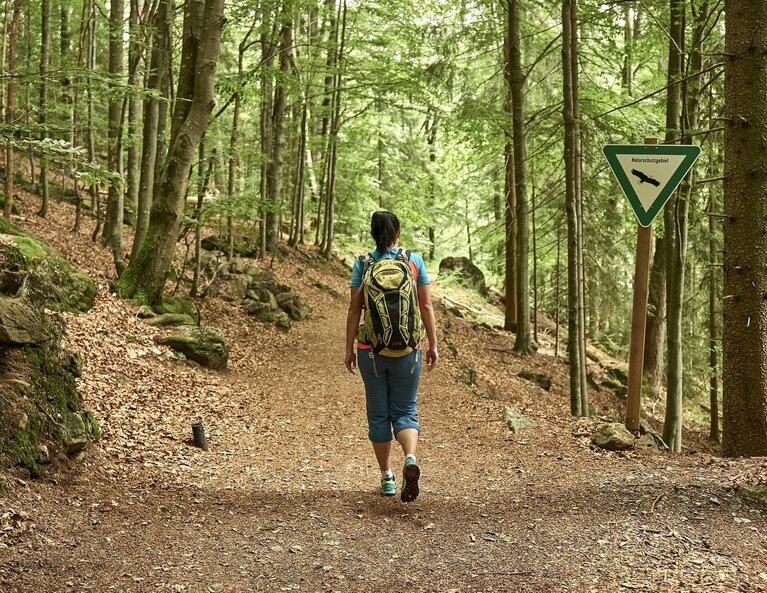 Eine Frau im Wanderoutfit geht auf einem Wanderweg durch den Wald. Am Rand des Weges steht ein dreieckiges Schild mit grünem Rand auf dem "Naturschutzgebiet" steht. | © Bodenmais Tourismus & Marketing GmbH