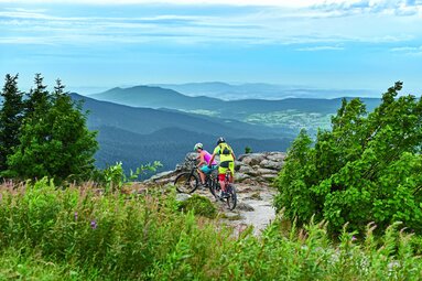 Zwei Radfahrer fahren auf einem Berg über einen Schotterweg. Hinter einem Felsen erkennt man den Bayerischen Wald mit seinen Gipfeln und Tälern. | © Bodenmais Tourismus & Marketing GmbH