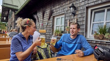 Ein Mann und eine Frau in sportlicher Kleidung sitzen in einem Biergarten und stoßen mit Maßkrügen voll Bier an. Auf dem Tisch vor ihnen liegt ein Fahrradhelm sowie eine Speisekarte, die verrät, dass sie sich in der Berghütte Schareben befinden. | © Bodenmais Tourismus & Marketing GmbH
