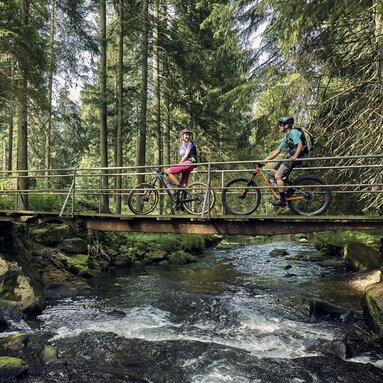 Eine Frau und ein Mann sitzen auf ihrem Rad. Sie befinden sich auf einer Brücke über einem Fluss in einem Waldstück. Sie halten sich am Brückengeländer fest und lachen einander an. | © Bodenmais Tourismus & Marketing GmbH