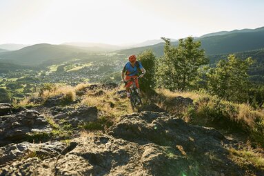 Ein Mann fährt mit dem Rad über steiniges Gelände. Er befindet sich auf einem Berg, hinter ihm sieht man das Tal liegen. Die Sonne geht hinter den Bergen des Bayerischen Waldes unter. | © Bodenmais Tourismus & Marketing GmbH