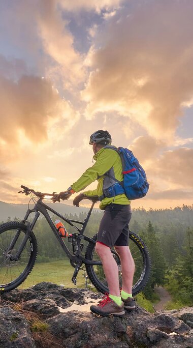 Ein Mann steht in Radkleidung auf einem Gipfel und hält sein Rad. Er steht mit dem Gesicht Richtung Sonnenuntergang, welchen den Himmel hinter ihm orange einfärbt. | © Bodenmais Tourismus & Marketing GmbH