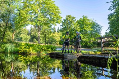 Eine Frau und ein Mann stehen an einem Weiher am Waldrand. Sowohl die beiden als auch die Natur spiegelt sich im Wasser. | © Bodenmais Tourismus & Marketing GmbH