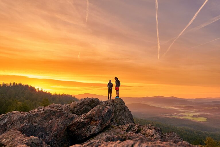 Eine Frau und ein Mann in sportlicher Kleidung stehen auf einem Berggipfel. Hinter ihnen geht die Sonne unter und der Himmel brennt förmlich. | © Bodenmais Tourismus & Marketing GmbH