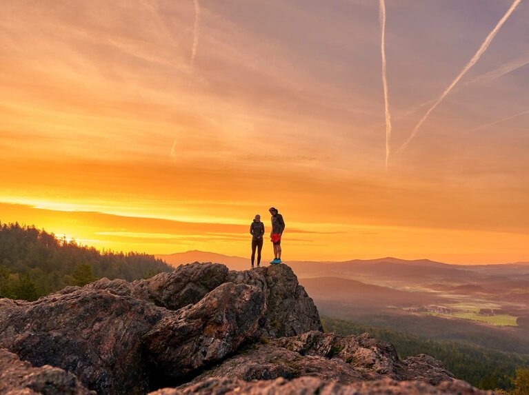Eine Frau und ein Mann in sportlicher Kleidung stehen auf einem Berggipfel. Hinter ihnen geht die Sonne unter und der Himmel brennt förmlich. | © Bodenmais Tourismus & Marketing GmbH