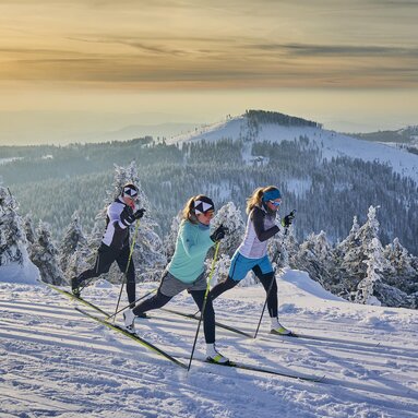 Drei Langläuferinnen fahren auf klassische Weise auf einer Loipe. Alles ist mit Schnee bedeckt und die untergehende Sonne färbt den Himmel orange. | © Bodenmais Tourismus & Marketing GmbH