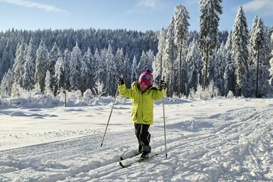 Ein kleines Mädchen steht auf Langlauf Skiern und hält Stöcke in der Hand. Sie fährt in der Spur und streckt vor Konzentration die Zunge an die obere Lippe. | © Bodenmais Tourismus & Marketing GmbH
