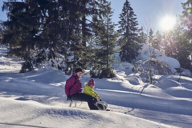 Eine Mutter fährt mit ihrem Kind, beide in Anorak und Schneehose, auf einem Schlitten auf einer verschneiten Piste den Hang hinunter. Neben der Piste stehen Bäume, durch die die Sonne scheint. | © Bodenmais Tourismus & Marketing GmbH
