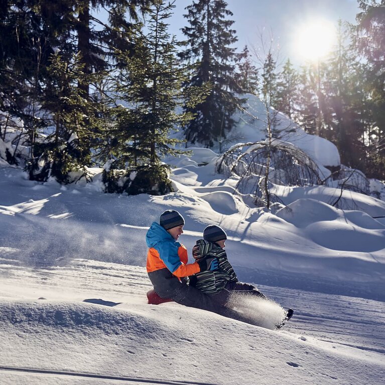 Zwei Jungen sitzen zusammen auf einem Schlitten und fahren deine verschneite Piste hinunter. Durch das Bremsen mit dem Fuß ist eine Schneewolke vor ihnen entstanden. | © Bodenmais Tourismus & Marketing GmbH