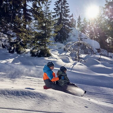 Zwei Jungen sitzen zusammen auf einem Schlitten und fahren deine verschneite Piste hinunter. Durch das Bremsen mit dem Fuß ist eine Schneewolke vor ihnen entstanden. | © Bodenmais Tourismus & Marketing GmbH