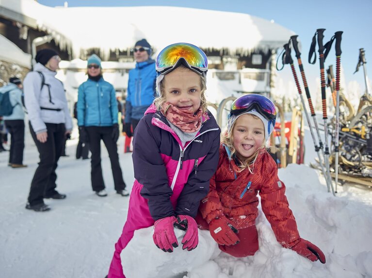 Zwei Mädchen in Winteranorak und Skibrille spielen auf einem Schneeberg. Neben ihnen stehen Skistöcke und mehrere Erwachsene. | © Bodenmais Tourismus & Marketing GmbH