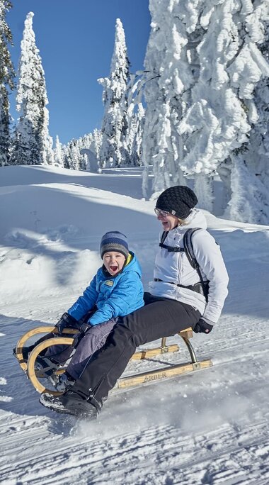 Mutter und Sohn sitzen zusammen auf einem Holzschlitten und fahren eine schneeweiße Piste hinunter. Vor strahlendblauem Himmel stehen schneebehangene Nadelbäume. | © Bodenmais Tourismus & Marketing GmbH