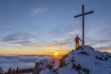 Ein Mann mit Schneeschuhen und Stöcken steht am Gipfelkreuz des Großen Arbers. Hinter ihm geht die Sonne unter und im Tal liegt schon der Nebel. | © Bodenmais Tourismus & Marketing GmbH