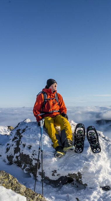Ein Mann in sportlicher Winterbekleidung sitzt auf einem verschneiten Berggipfel und hat Schneeschuhe neben sich liegen.  Er schaut in die Ferne. | © Bodenmais Tourismus & Marketing GmbH