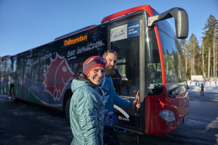 Zwei Winterwanderer stehen an einer Haltestelle vor einem roten Bus mit der Aufschrift "Ostbayernbus" | © Bodenmais Tourismus & Marketing GmbH