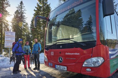 Drei Wanderer stehen an einer Bushaltestelle und halten Schneeschuhe und Wanderstöcke in der Hand. Rechts von ihnen fährt der Ostbayernbus ein. | © Bodenmais Tourismus & Marketing GmbH