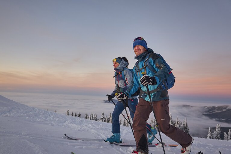 Zwei Skitourengeher sind bei Sonnenuntergang am Großen Arber. Der Himmel färbt sich lila und im Tal liegt der Nebel. | © Bodenmais Tourismus & Marketing GmbH