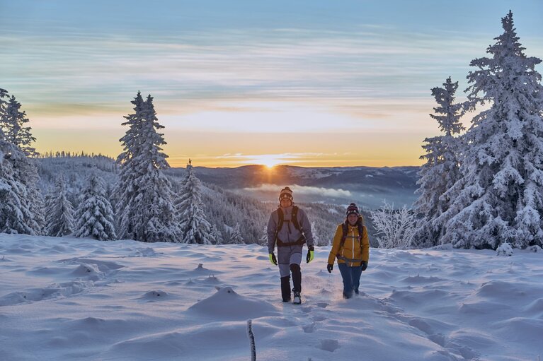 Eine Frau und ein Mann wandern durch den Schnee, der ihnen bis zur Mitte des Schienbeins reicht. Hinter ihnen verschwindet die Sonne hinter einem anderen Gipfel. | © Bodenmais Tourismus & Marketing GmbH