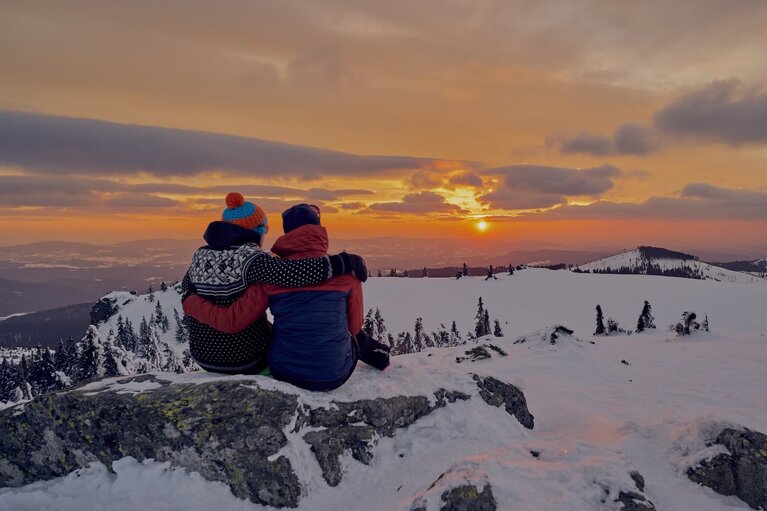 Zwei Personen sitzen zusammen auf einem Fels am Berggipfel, haben die Arme umeinander gelegt und schauen sich den Sonnenuntergang an. | © Bodenmais Tourismus & Marketing GmbH
