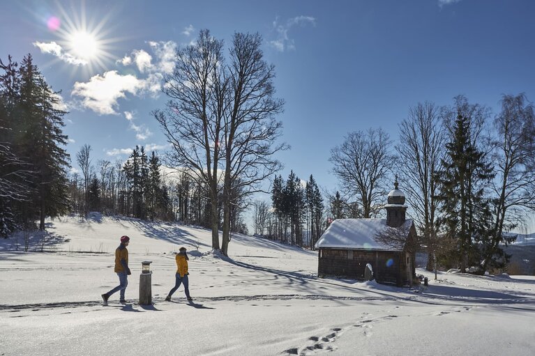 Zwei Personen wandern am verschneiten Harlachberg in Richtung der Kapelle der GutsAlm. | © Bodenmais Tourismus & Marketing GmbH