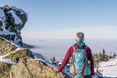 Eine Frau sitzt mit einem Rucksack im Bodenmais Design und Thermosflasche auf einem verschneiten Berggipfel. Sie blickt in die Ferne, wo der Nebel das Tal bedeckt und einzelne Berggipfel hervorschauen. | © Bodenmais Tourismus & Marketing GmbH