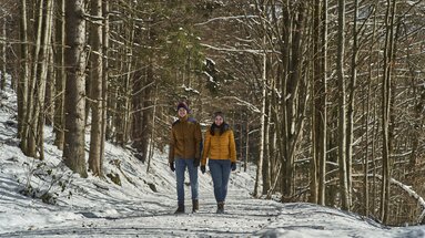 Eine Paar läuft auf einem breiten präpariertem Winterwanderweg durch den Wald. Auf dem Schnee liegen vereinzelt Blätter. | © Bodenmais Tourismus & Marketing GmbH