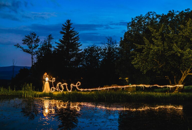 Bei Dämmerung küsst sich ein Brautpaar neben dem Teich der GutsAlm Harlachberg stehend. Durch Dauerbelichtung wird der Schriftzug "love" aus Licht sichtbar. | © jokumaxx design