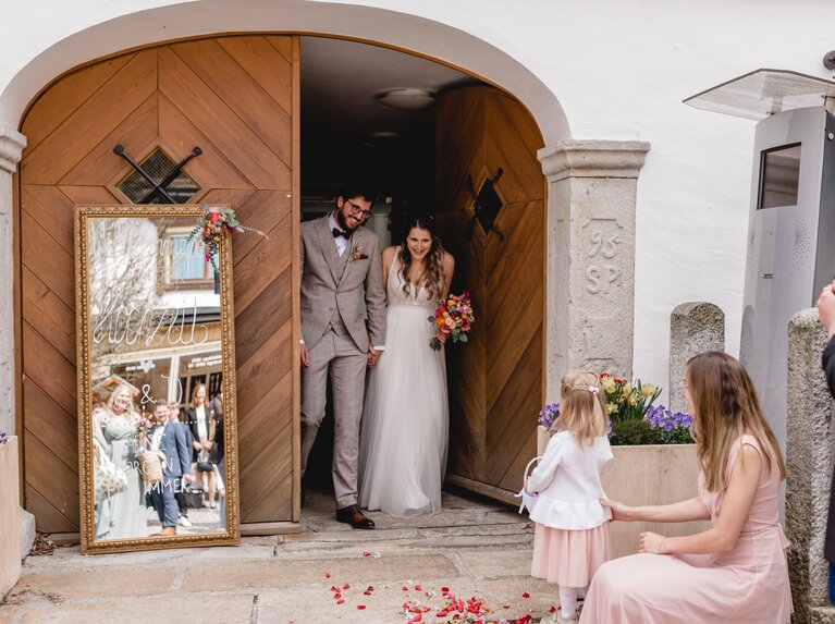 Das Brautpaar kommt aus dem Tor des Alten Rathauses vor dem Blütenblätter liegen und die Gäste warten. | © Lisa Bauer Photography