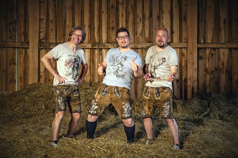 Drei Männer mit Lederhose stehen in einem Stall mit Heu. | © Out of Bayern