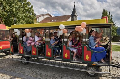 In einer Bimmelbahn sitzen Hochzeitsgäste mit Luftballoons | © Lisa Bauer Photography