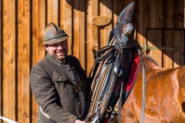 Ein Mann mit Trachtenjacke und Hut hält ein Kumt hoch um die Pferde vor die Kutsche zu spannen. | © Bodenmais Tourismus & Marketing GmbH