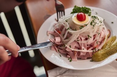 Auf einem Tisch steht ein Teller Wurstsalat. Dieser ist garniert mit feingeschnittenen weißen Zwiebeln, einer Scheibe gekochtem Ei, einer Cocktailtomate sowie Petersilie und einer Essiggurke.  | © Bodenmais Tourismus & Marketing GmbH