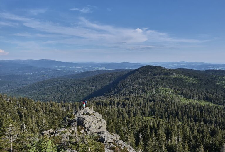 Ein Paar steht auf einem Felsvorsprung. Da sie ziemlich weit weg stehen erkennt man sie nur an ihren bunten T-shirts. Sie blicken über die mit Wald bewachsenen Berge des Bayerischen Waldes. | © Bodenmais Tourismus & Marketing GmbH