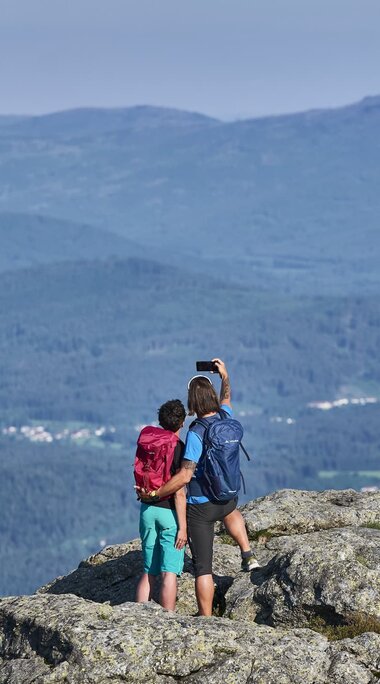 Ein Paar steht auf einem Gipfel und macht ein Selfie. Dabei haben Sie einen wunderbaren Blick in die Ferne. | © Bodenmais Tourismus & Marketing GmbH